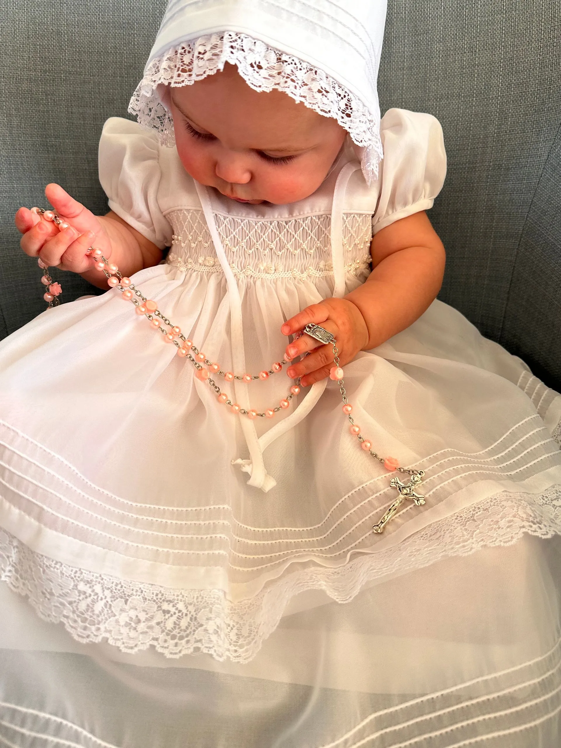 A baby in a white dress is holding a rosary