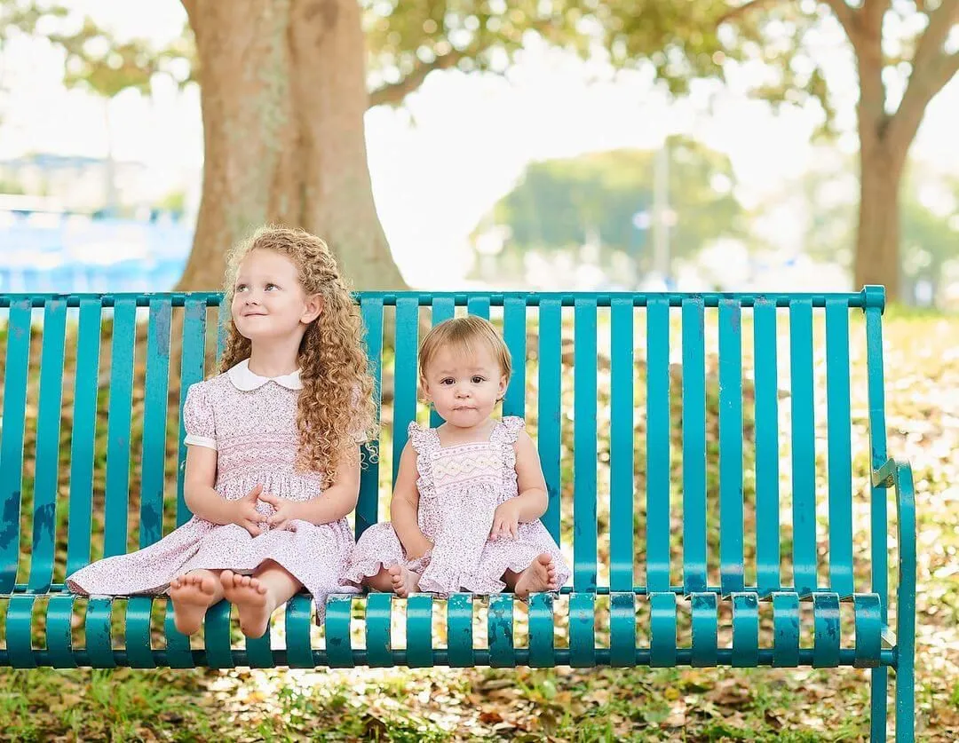 Two little girls are sitting on a blue bench in a park.